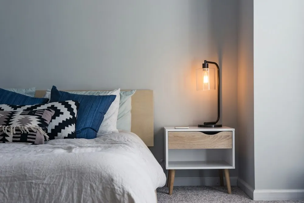 Bedside corner lit by a warm pendant lamp, showing low-level layered lighting against a pale wall.